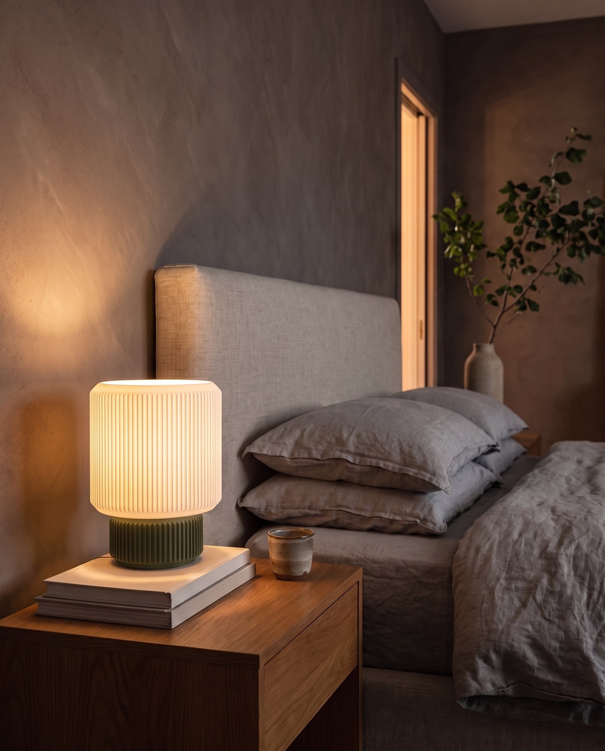 Lámpara Ágora con base Musgo, encendida en una mesa de noche junto a la cama